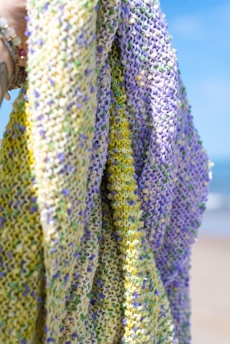 A multi-colored knitted fabric being held up, with a mix of yellow, purple, and green yarn. The knitted texture is prominently visible, and the background appears to be a beach with a clear blue sky and some sea visible.