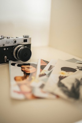 A vintage camera is placed on a light-colored surface next to several printed photos of portraits. The photos are slightly overlapping, indicating a casual arrangement.