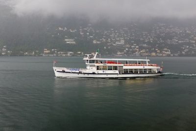 A ferry sails across a calm body of water with a misty hillside in the background. The ferry displays a flag at the stern and is transporting numerous passengers whose seats are covered with red cushions. Small houses dot the hillside under foggy clouds.