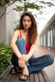 A woman with long brown hair is sitting on a wooden bench, wearing a red sleeveless top and blue jeans. She appears calm and composed, with her legs crossed and hands resting on her knee. The background features greenery and a building with large windows, suggesting an outdoor setting.