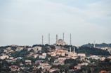 A scenic view of a cityscape with a large, prominent mosque featuring multiple minarets atop a hill. The foreground is filled with densely packed residential buildings, surrounded by lush green trees. The sky above is clear with a light blue hue, adding to the serene atmosphere.