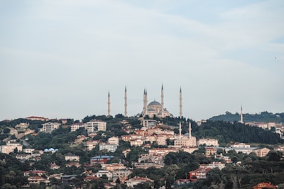 A scenic view of a cityscape with a large, prominent mosque featuring multiple minarets atop a hill. The foreground is filled with densely packed residential buildings, surrounded by lush green trees. The sky above is clear with a light blue hue, adding to the serene atmosphere.