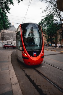 A modern red-and-white tram is moving along tracks on a city street with pedestrians in the background. The tram has a sleek design with large windows and is surrounded by an urban setting featuring buildings, trees, and parked cars.