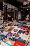 A bookstore or book market with a variety of books displayed on tables. Books of different genres and languages can be seen stacked closely together. A person is sitting in the background, possibly reading or using a phone, amidst shelves filled with more books.