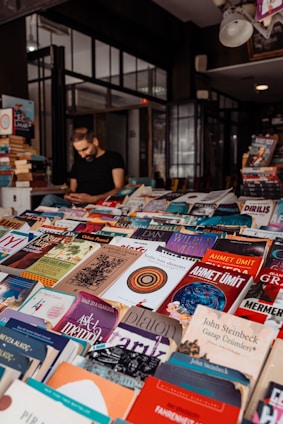 A bookstore or book market with a variety of books displayed on tables. Books of different genres and languages can be seen stacked closely together. A person is sitting in the background, possibly reading or using a phone, amidst shelves filled with more books.