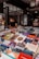 A bookstore or book market with a variety of books displayed on tables. Books of different genres and languages can be seen stacked closely together. A person is sitting in the background, possibly reading or using a phone, amidst shelves filled with more books.