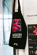 A black tote bag hangs on a wall featuring a colorful abstract design of red and purple shapes. The text on the bag reads 'DeSIGN DEPARTMENT' with additional information at the bottom. The background includes part of a white poster with text and a partial view of a window.