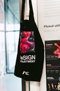 A black tote bag hangs on a wall featuring a colorful abstract design of red and purple shapes. The text on the bag reads 'DeSIGN DEPARTMENT' with additional information at the bottom. The background includes part of a white poster with text and a partial view of a window.