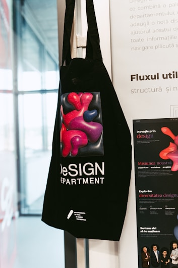 A black tote bag hangs on a wall featuring a colorful abstract design of red and purple shapes. The text on the bag reads 'DeSIGN DEPARTMENT' with additional information at the bottom. The background includes part of a white poster with text and a partial view of a window.
