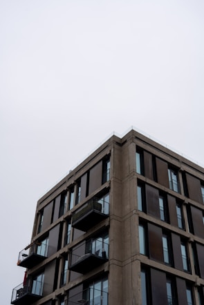 A modern apartment building with a minimalist design is depicted, featuring multiple floors with large glass windows and small balconies. The sky is overcast, giving a muted backdrop to the structure.