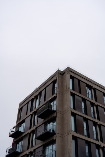 A modern apartment building with a minimalist design is depicted, featuring multiple floors with large glass windows and small balconies. The sky is overcast, giving a muted backdrop to the structure.