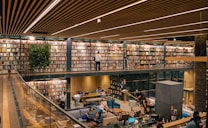 A modern library with two levels, featuring a vast collection of books neatly arranged on shelves. The upper level includes a walkway with individuals browsing the books. The lower level is a cozy seating area with people engaged in reading and conversation. The architecture includes wooden panels on the ceiling, glass railings, and plants adding a touch of greenery.