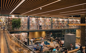 A modern library with two levels, featuring a vast collection of books neatly arranged on shelves. The upper level includes a walkway with individuals browsing the books. The lower level is a cozy seating area with people engaged in reading and conversation. The architecture includes wooden panels on the ceiling, glass railings, and plants adding a touch of greenery.