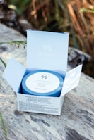 A Dr.Ceuracle Hyal Reyouth Hydrogel Eye Mask package is open on a wooden surface. The blue and white packaging is set against a blurred background of natural foliage, with sunlight gently illuminating the product.