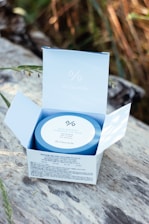 A Dr.Ceuracle Hyal Reyouth Hydrogel Eye Mask package is open on a wooden surface. The blue and white packaging is set against a blurred background of natural foliage, with sunlight gently illuminating the product.