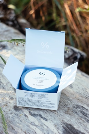 A Dr.Ceuracle Hyal Reyouth Hydrogel Eye Mask package is open on a wooden surface. The blue and white packaging is set against a blurred background of natural foliage, with sunlight gently illuminating the product.