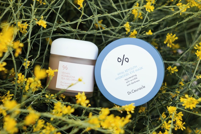 Two skincare products are placed amidst yellow blossoming flowers with green stems. The first product has a round blue lid labeled 'Hyal Reyouth Hydrogel Eye Mask' and the second is a jar of 'Vegan Kombucha Tea Gel Cream'.