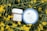 Two skincare products are placed amidst yellow blossoming flowers with green stems. The first product has a round blue lid labeled 'Hyal Reyouth Hydrogel Eye Mask' and the second is a jar of 'Vegan Kombucha Tea Gel Cream'.
