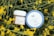 Two skincare products are placed amidst yellow blossoming flowers with green stems. The first product has a round blue lid labeled 'Hyal Reyouth Hydrogel Eye Mask' and the second is a jar of 'Vegan Kombucha Tea Gel Cream'.