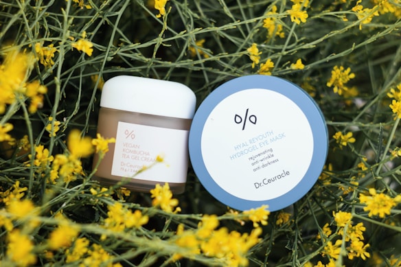 Two skincare products are placed amidst yellow blossoming flowers with green stems. The first product has a round blue lid labeled 'Hyal Reyouth Hydrogel Eye Mask' and the second is a jar of 'Vegan Kombucha Tea Gel Cream'.