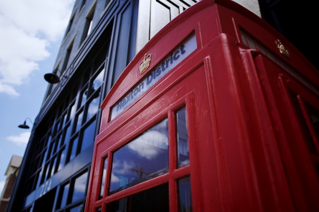 A classic red British telephone booth is prominently featured against a backdrop of modern black and glass architecture. The booth is labeled with 'Brixton District' and reflects the surrounding environment in its windows. The sky is partly cloudy.