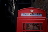 A red telephone booth with the sign 'Haxton District' and the phrase 'Where Art & Business Collide'. The booth features a crown emblem above the sign. The background includes a brick wall and a dark doorway, creating a contrast with the vibrant red of the booth.