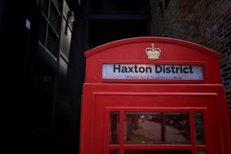 A red telephone booth with the sign 'Haxton District' and the phrase 'Where Art & Business Collide'. The booth features a crown emblem above the sign. The background includes a brick wall and a dark doorway, creating a contrast with the vibrant red of the booth.