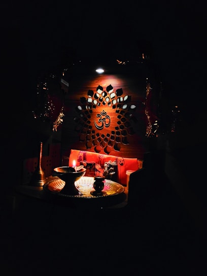 An intricately designed wooden panel with a central Om symbol is illuminated softly. Below, a small altar setup includes a lit oil lamp, a brass plate, and various religious artifacts, creating a serene and spiritual atmosphere.