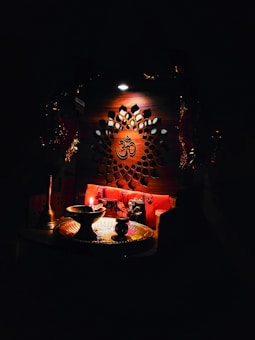 An intricately designed wooden panel with a central Om symbol is illuminated softly. Below, a small altar setup includes a lit oil lamp, a brass plate, and various religious artifacts, creating a serene and spiritual atmosphere.