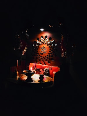 An intricately designed wooden panel with a central Om symbol is illuminated softly. Below, a small altar setup includes a lit oil lamp, a brass plate, and various religious artifacts, creating a serene and spiritual atmosphere.