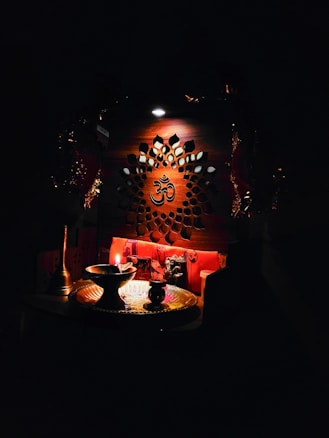 An intricately designed wooden panel with a central Om symbol is illuminated softly. Below, a small altar setup includes a lit oil lamp, a brass plate, and various religious artifacts, creating a serene and spiritual atmosphere.