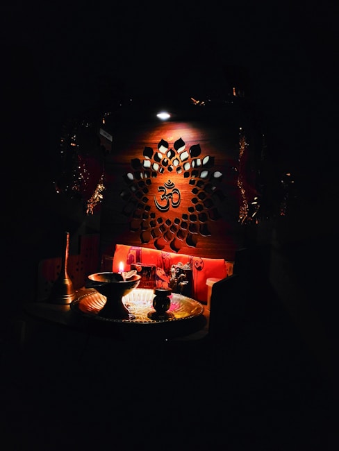 An intricately designed wooden panel with a central Om symbol is illuminated softly. Below, a small altar setup includes a lit oil lamp, a brass plate, and various religious artifacts, creating a serene and spiritual atmosphere.