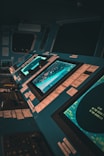 A control room with multiple large monitors displaying various data and maps. The environment has a modern and high-tech feel, with dim lighting creating a focused atmosphere. The screens are surrounded by numerous buttons and switches.