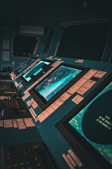 A control room with multiple large monitors displaying various data and maps. The environment has a modern and high-tech feel, with dim lighting creating a focused atmosphere. The screens are surrounded by numerous buttons and switches.
