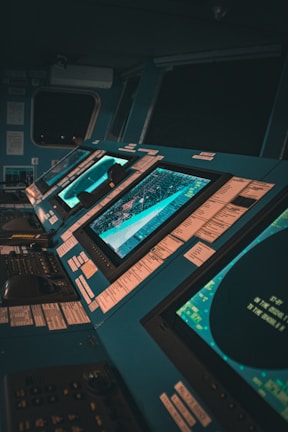 A control room with multiple large monitors displaying various data and maps. The environment has a modern and high-tech feel, with dim lighting creating a focused atmosphere. The screens are surrounded by numerous buttons and switches.