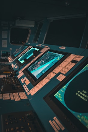 A control room with multiple large monitors displaying various data and maps. The environment has a modern and high-tech feel, with dim lighting creating a focused atmosphere. The screens are surrounded by numerous buttons and switches.