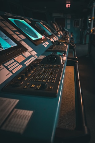A dimly lit control room featuring several computer monitors and control panels, with buttons and knobs. The space appears to be high-tech, possibly in a naval or air traffic setting.