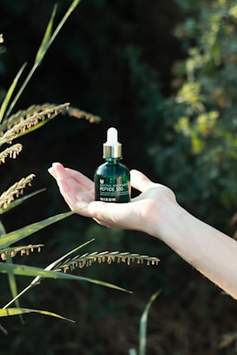 A hand is holding a small green bottle with a dropper cap, surrounded by green foliage in a natural, outdoor setting. The text on the bottle reads 'Original Skin Energy Peptide 500' from the brand 'Mizon'.