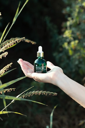 A hand is holding a small green bottle with a dropper cap, surrounded by green foliage in a natural, outdoor setting. The text on the bottle reads 'Original Skin Energy Peptide 500' from the brand 'Mizon'.