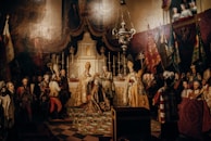 A historical painting depicts a grand religious ceremony with elaborate attire and regalia. Clergy members are in ornate robes, surrounded by nobility and officials. The setting is opulent with a richly decorated altar, candles, and luxurious drapery. People are gathered solemnly, indicating a significant event.