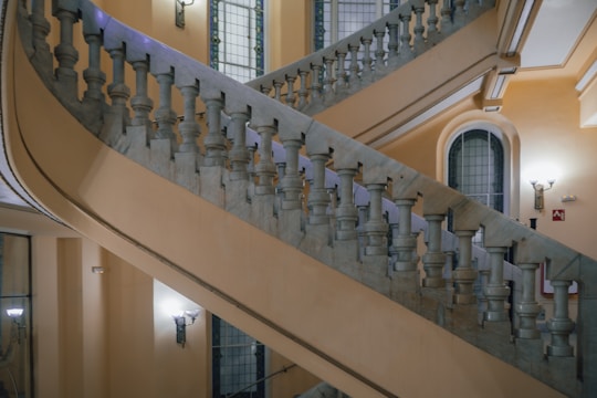 An ornate, multi-level staircase with marble balustrades, set against a backdrop of softly lit walls. The stairwell is complemented by arched windows with decorative glass and iron wall sconces.