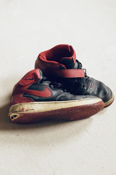 A pair of worn black and red high-top sneakers is placed on a light-colored surface. The shoes, featuring red accents and a prominent brand logo, display signs of wear with scuffed soles and creased leather.
