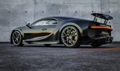 A sleek, high-performance sports car with a dark metallic finish is parked on a textured surface in front of a smooth concrete wall. The vehicle features aerodynamic designs with a prominent rear spoiler, large alloy wheels, and a low profile that suggests speed and luxury.