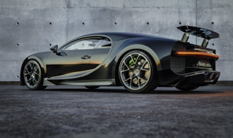 A sleek, high-performance sports car with a dark metallic finish is parked on a textured surface in front of a smooth concrete wall. The vehicle features aerodynamic designs with a prominent rear spoiler, large alloy wheels, and a low profile that suggests speed and luxury.