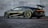 A sleek, high-performance sports car with a dark metallic finish is parked on a textured surface in front of a smooth concrete wall. The vehicle features aerodynamic designs with a prominent rear spoiler, large alloy wheels, and a low profile that suggests speed and luxury.