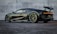 A sleek, high-performance sports car with a dark metallic finish is parked on a textured surface in front of a smooth concrete wall. The vehicle features aerodynamic designs with a prominent rear spoiler, large alloy wheels, and a low profile that suggests speed and luxury.