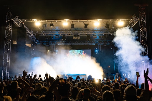 A crowded concert venue with a large stage, bright lights, and smoke effects, featuring a lively audience with raised hands and phones capturing the moment.
