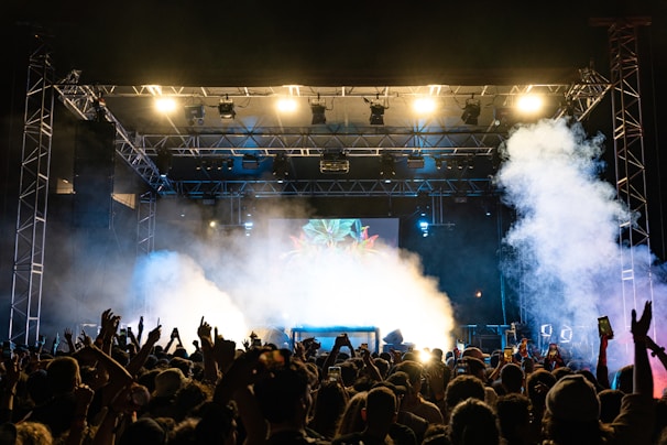 A crowded concert venue with a large stage, bright lights, and smoke effects, featuring a lively audience with raised hands and phones capturing the moment.