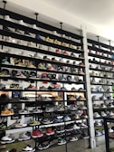 A shoe store wall filled with multiple rows of sneakers, displaying a variety of brands, styles, and colors. The sneakers are arranged neatly on black shelves against a white wall. There are hints of other merchandise and clothing visible in the background.