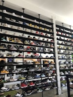 A shoe store wall filled with multiple rows of sneakers, displaying a variety of brands, styles, and colors. The sneakers are arranged neatly on black shelves against a white wall. There are hints of other merchandise and clothing visible in the background.